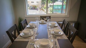 A dining table set for six with white plates, bowls, and clear glasses, positioned near a window with a view of a residential area.