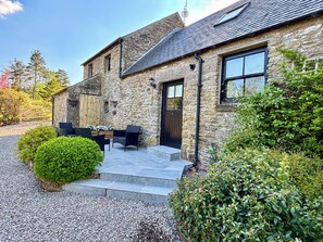 Un bâtiment en pierre avec une porte noire, une terrasse dotée de mobilier d’extérieur et une allée gravillonnée.