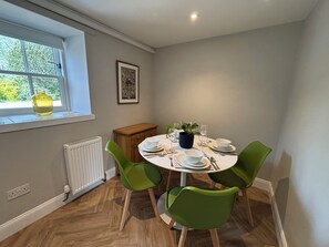 Un coin repas avec une table ronde, des chaises vertes, une plante en pot et une fenêtre donnant sur des arbres.