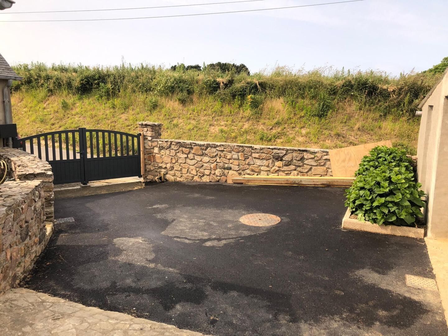 A gated driveway leading to a stone wall, a manhole cover, and a small green bush.
