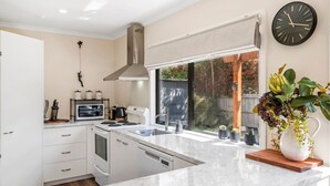 Una cocina moderna con armarios blancos, una campana extractora de acero inoxidable, un horno empotrado y un amplio ventanal con vistas a una terraza de madera y árboles.