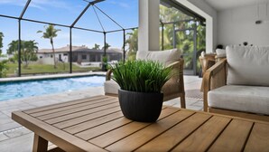 A poolside area with a wooden table, a black pot with green plants, a white sofa, and a ceiling fan. - Paradise in front of the water (Cape Coral)