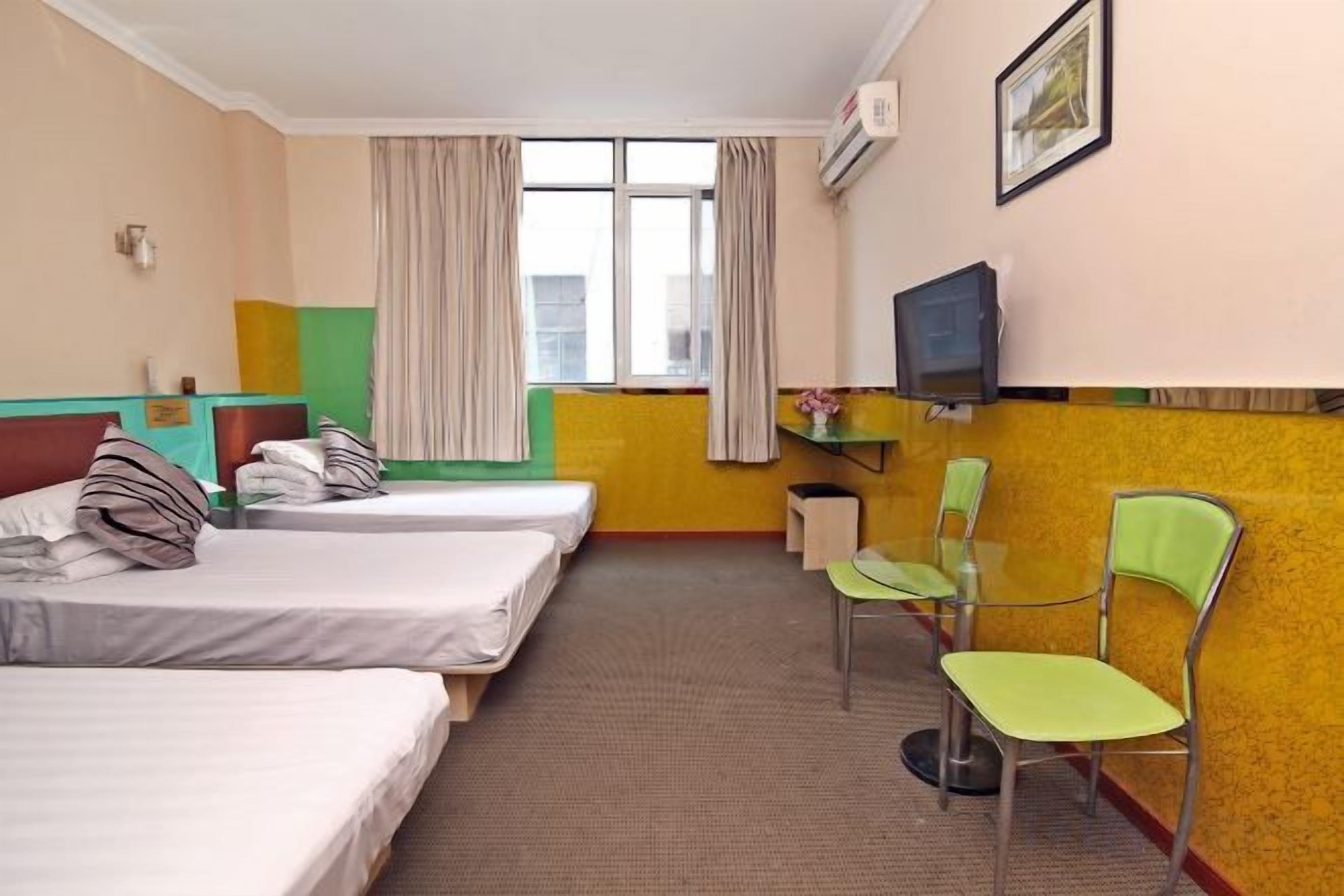 Quarto de hotel com três camas de solteiro, uma mesa com cadeira, televisão e janela com cortinas.