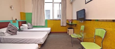 Quarto de hotel com três camas de solteiro, uma mesa com cadeira, televisão e janela com cortinas.
