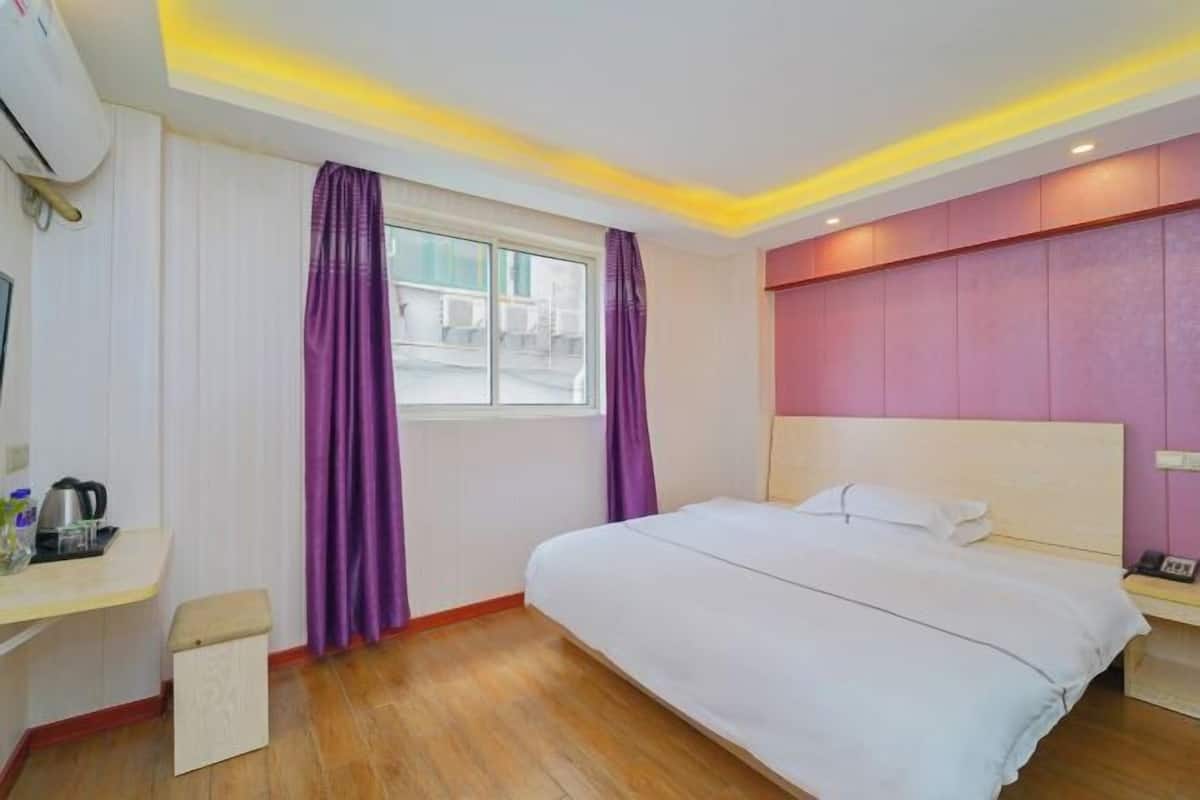 A hotel room with a bed, a desk, a window with purple curtains, and a wall with a pink panel.