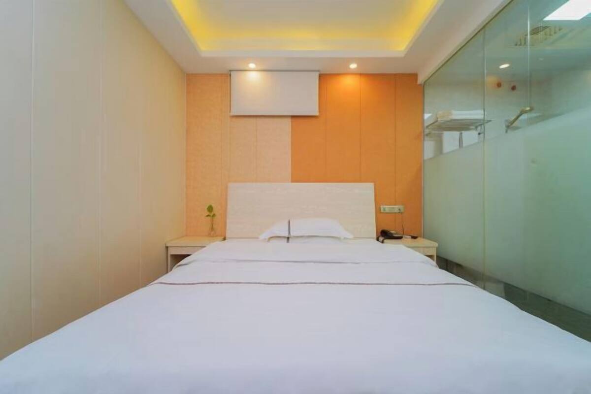 A hotel room with a large bed, a bedside table with a lamp, a wall-mounted TV, and a glass shower enclosure.