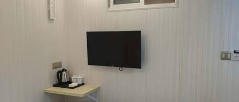 A hotel room with a bed, a TV mounted on the wall, a small table with a kettle and cups, and a window.
