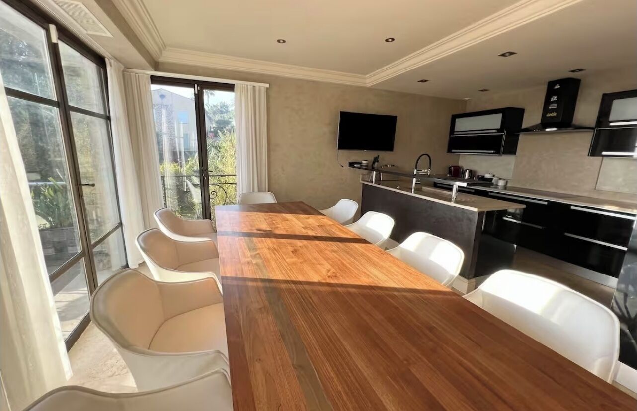 Eine moderne Küche mit einem Holzesstisch und weißen Stühlen, einem über der Arbeitsfläche montierten Fernseher und einem Balkon mit Blick auf Grünflächen.