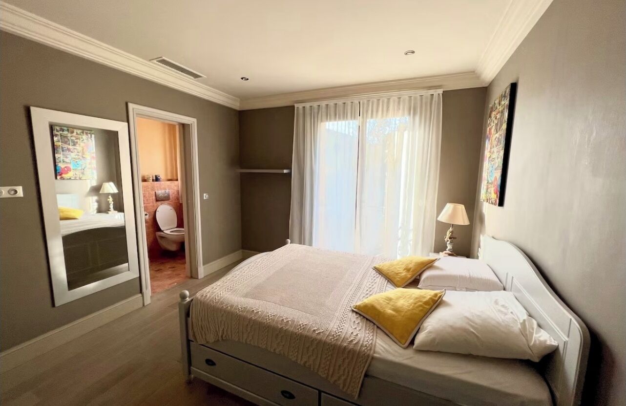 A bedroom with a bed, a bedside table, a window with curtains, and a bathroom visible through an open door.