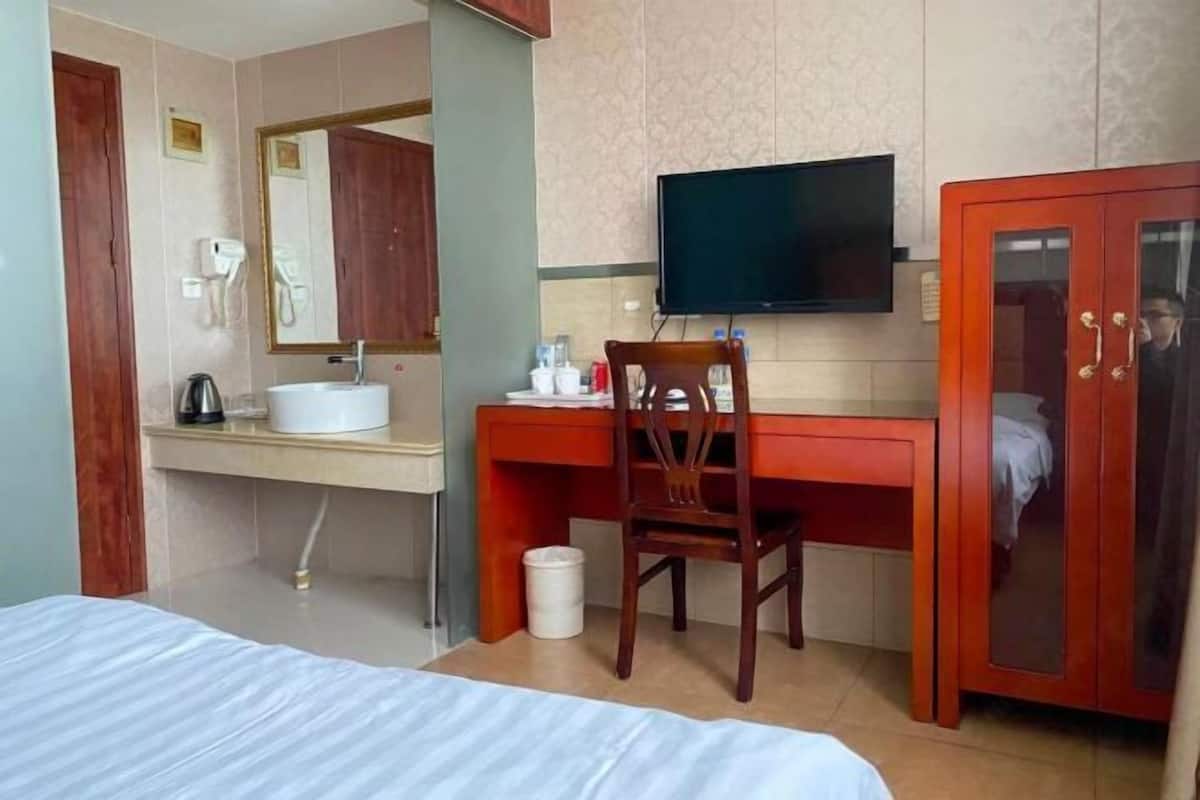 Quarto de hotel com cama, escrivaninha com cadeira, televisão, armário e banheiro com pia e espelho.