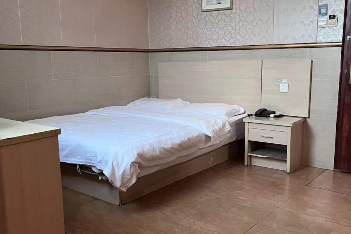 Uma cama de solteiro com cabeceira de madeira, uma mesinha de cabeceira com abajur e uma tomada na parede.