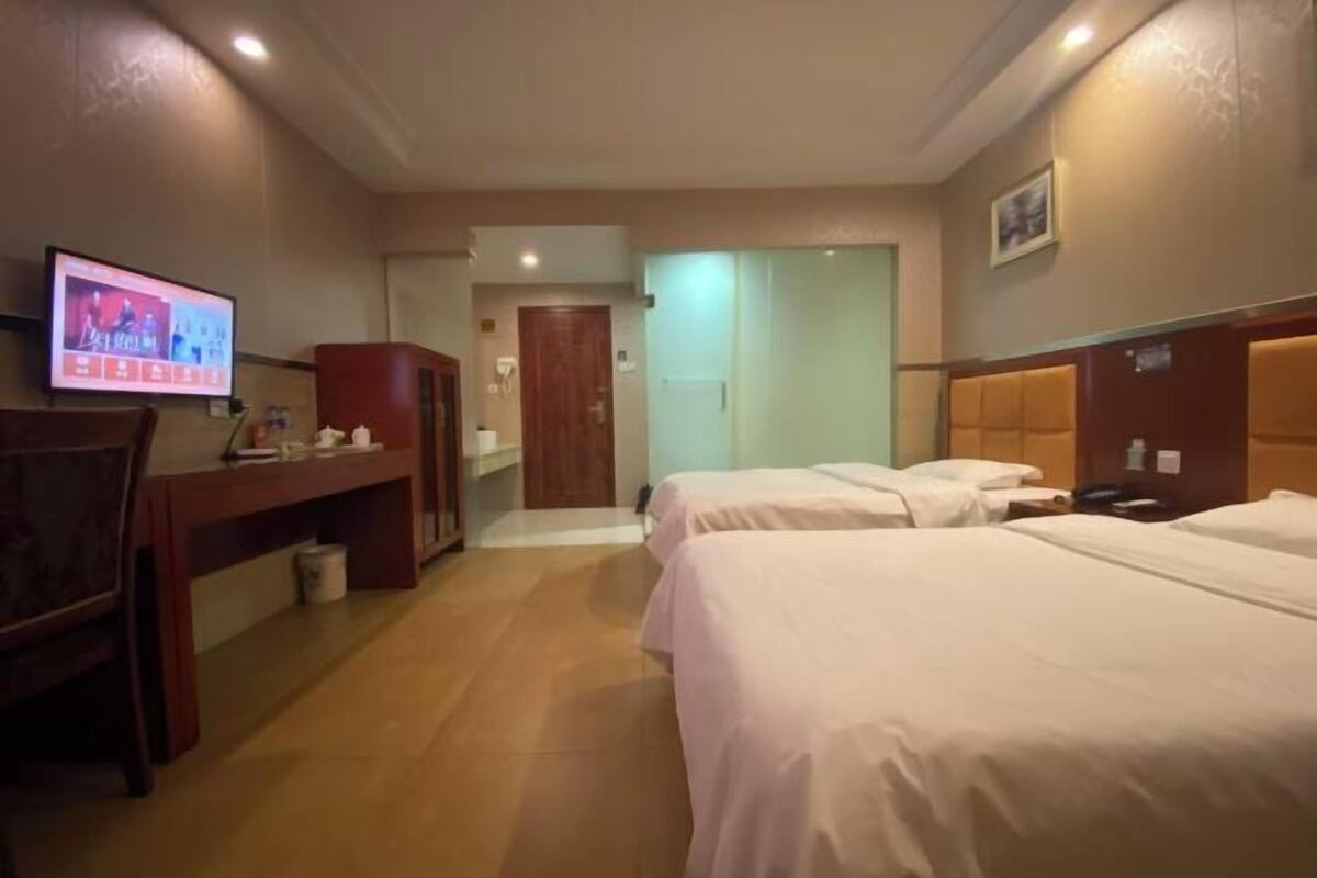 Quarto de hotel com duas camas, uma escrivaninha, uma televisão e uma porta que dá para outro cômodo.