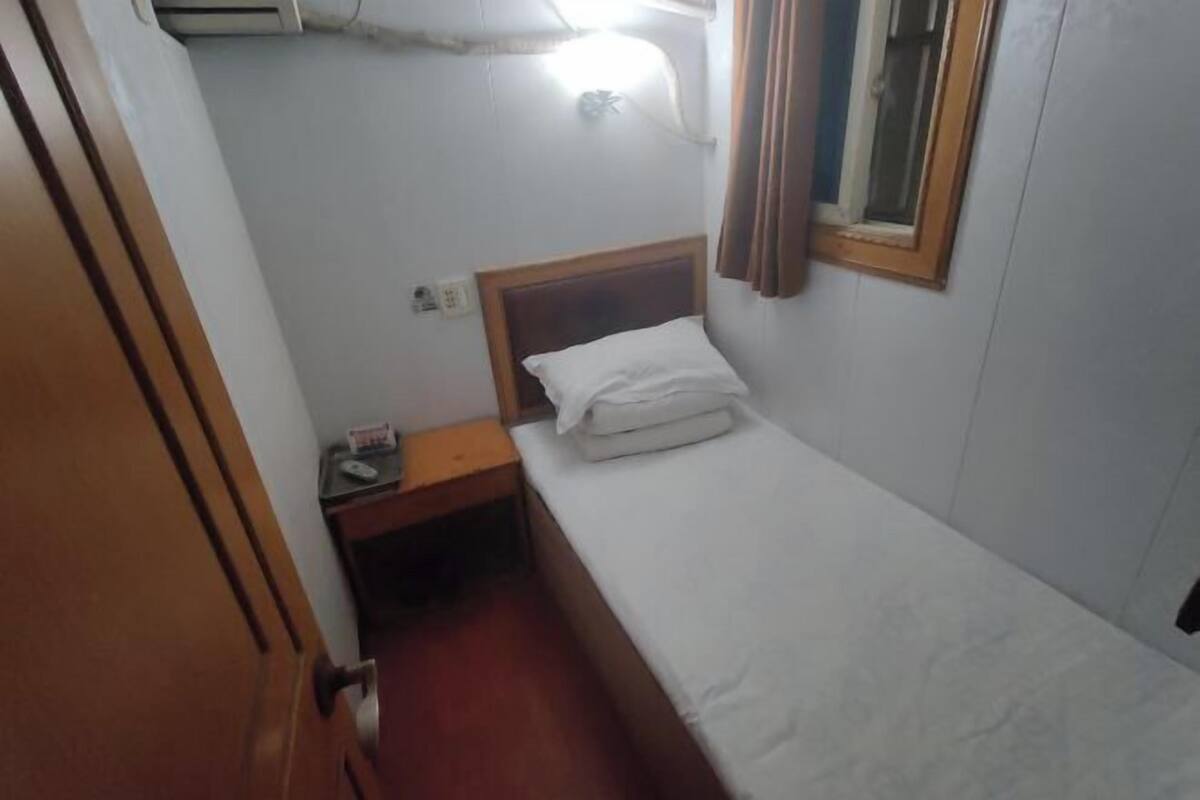 A small, single-bed room with a wooden headboard, a bedside table, and a window with curtains.
