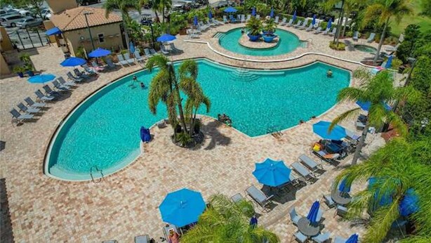 A large, curved swimming pool with lounge chairs and umbrellas, surrounded by palm trees and a tiled deck. - Resort Style 3 Bed/2 Bath Garage Home in GATED River Strand Golf & Country Club! (Bradenton)
