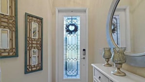 A hallway with a white door featuring a decorative glass panel and a wreath. There are two wall-mounted decorative panels and a round mirror above a white dresser with two ornate vases. - Resort Style 3 Bed/2 Bath Garage Home in GATED River Strand Golf & Country Club! (Bradenton)