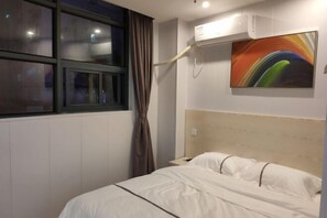 Una habitación de hotel con cama, ventana con cortinas, equipo de aire acondicionado y un cuadro abstracto de colores en la pared.