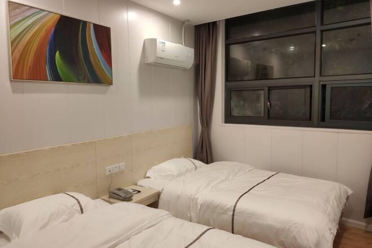 A hotel room with two beds, a wall-mounted air conditioner, a window with curtains, and a colorful abstract painting.