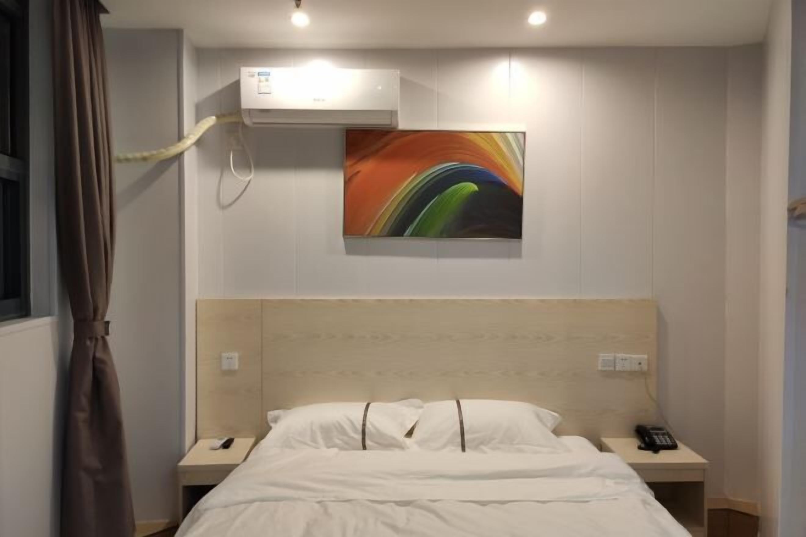 A hotel room with a bed, bedside tables, a phone, an air conditioning unit, and a colorful abstract painting on the wall.