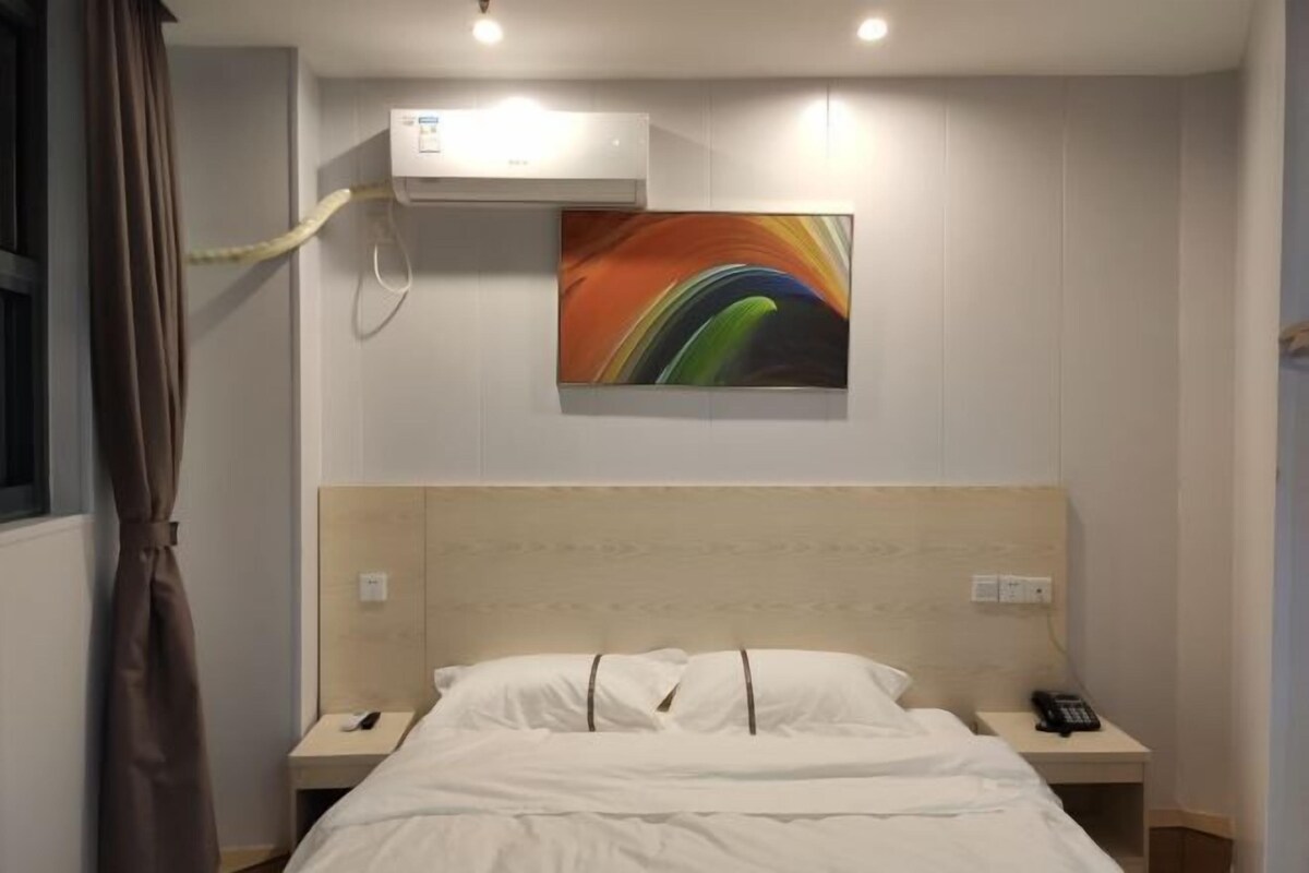 A hotel room with a bed, bedside tables, a phone, an air conditioning unit, and a colorful abstract painting on the wall.