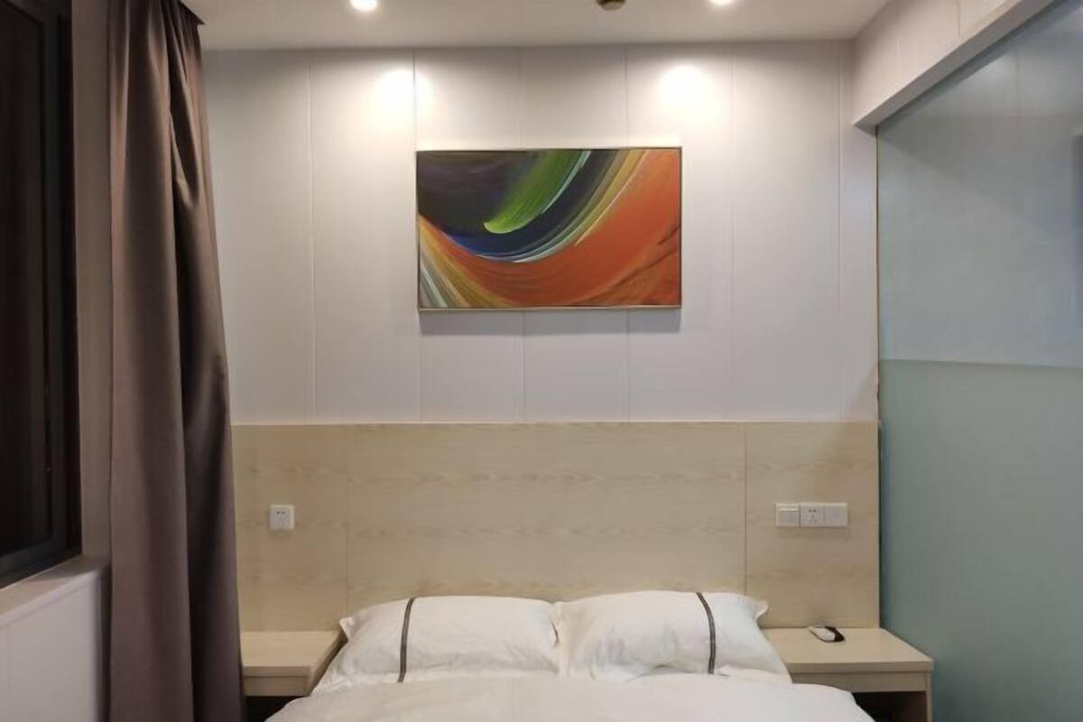 A hotel room with a bed, a nightstand, a wall-mounted abstract painting, and a window with curtains.