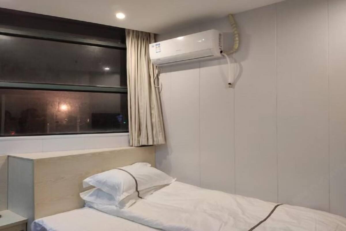 A hotel room with a single bed, a window with curtains, an air conditioning unit, and a bedside table.