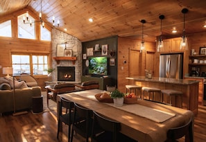 A cozy cabin interior with a stone fireplace, wooden dining area, and a kitchen with a refrigerator and bar stools.