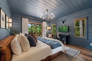 A bedroom with a large bed, a desk, and a chair. There are windows with curtains, a chandelier, and a mounted deer head on the wall.
