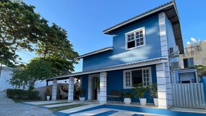 Une maison à deux étages, à l’extérieur bleu et aux ornements blancs, dotée d’un patio couvert agrémenté de plantes en pot et d’un hamac.