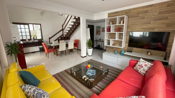 Un salon moderne avec un canapé rouge, un canapé jaune et une table basse en verre. On y trouve un escalier menant à un autre niveau, un coin repas avec des chaises et un téléviseur fixé à un mur en bois.