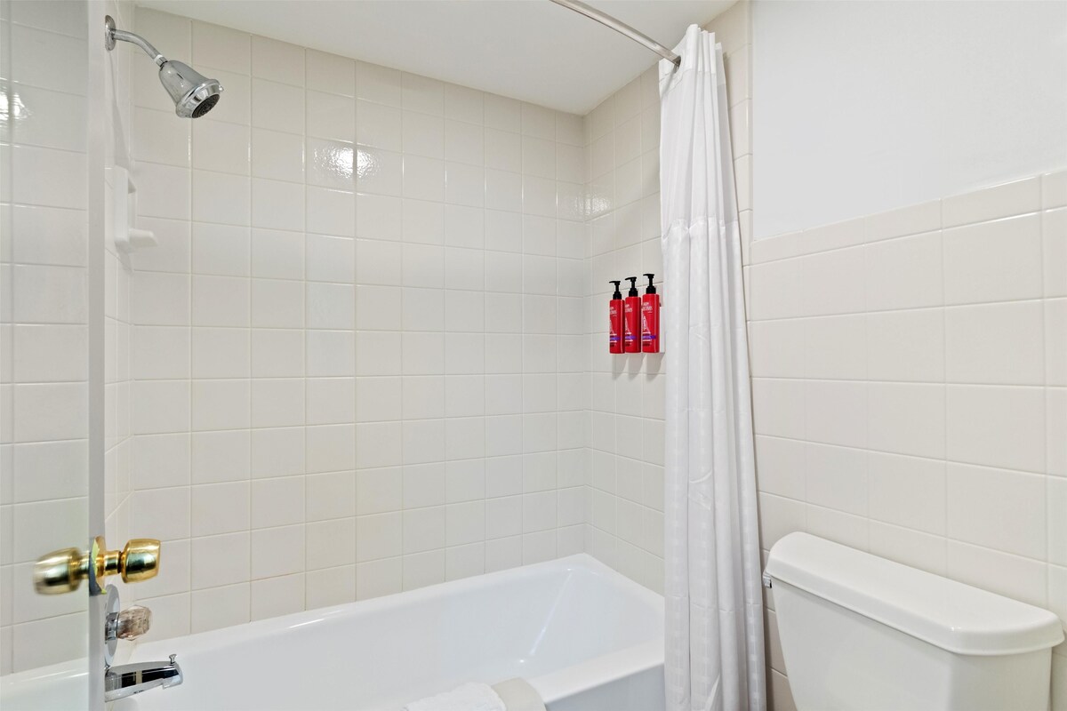 Een badkamer met een witte badkuip, een douchekop en een toilet. Er staan drie rode flessen aan de muur.