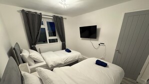 Ein Hotelzimmer mit zwei Betten, weißer Bettwäsche, grauen Kopfteilen, einem flachbildfernseher an der Wand und einer Tür mit grauem Paneel.