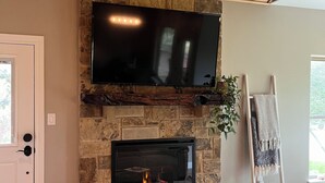 A living room with a stone fireplace, a mounted TV, and a wooden ceiling. - Cosy 1-bedroom cottage with spring-fed creek and spectacular views (Kerrville)