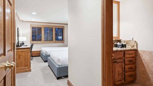 A hotel room with two beds, a desk, a chair, a window with blinds, a mirror, and a bathroom with a vanity and sink.