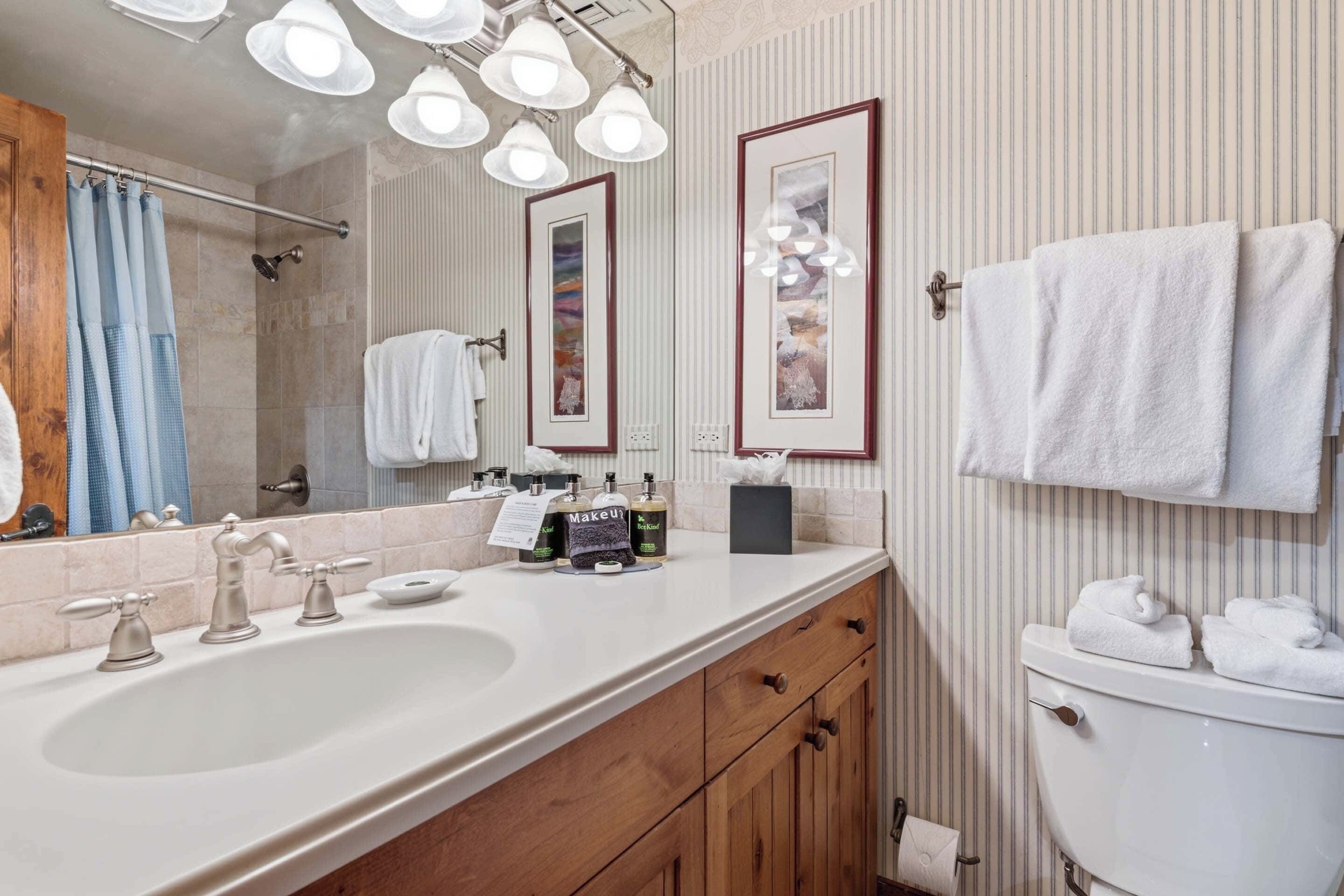 A bathroom with a large sink, vanity, mirror, and toilet. There are multiple light fixtures above the mirror and a towel rack with towels.