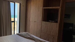 A bedroom with a bed, a wooden wardrobe, a television, and a view of the beach.