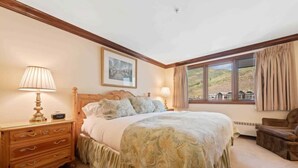 Une chambre à coucher avec un grand lit, des tables de chevet en bois, une commode et une fenêtre donnant sur des collines.