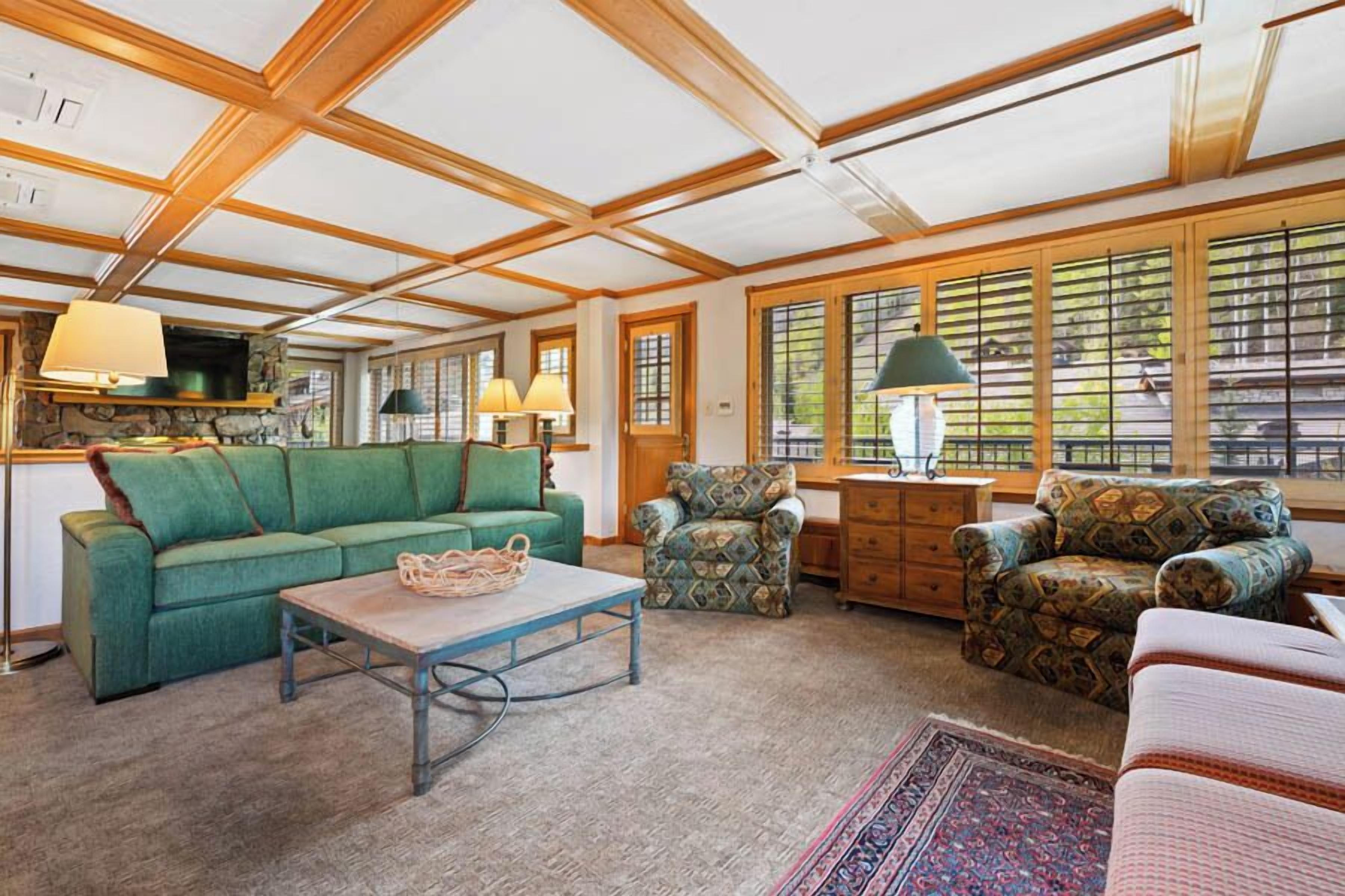 A spacious living room with a high ceiling, wooden beams, a green sofa, patterned armchairs, a coffee table, and a large window with blinds.