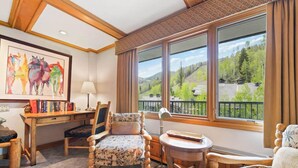 A room with a wooden desk, two chairs, a bed, and large windows offering a view of a forested landscape. - Lodge Tower 599 1BR  at the base of Vail Mountain (Vail)