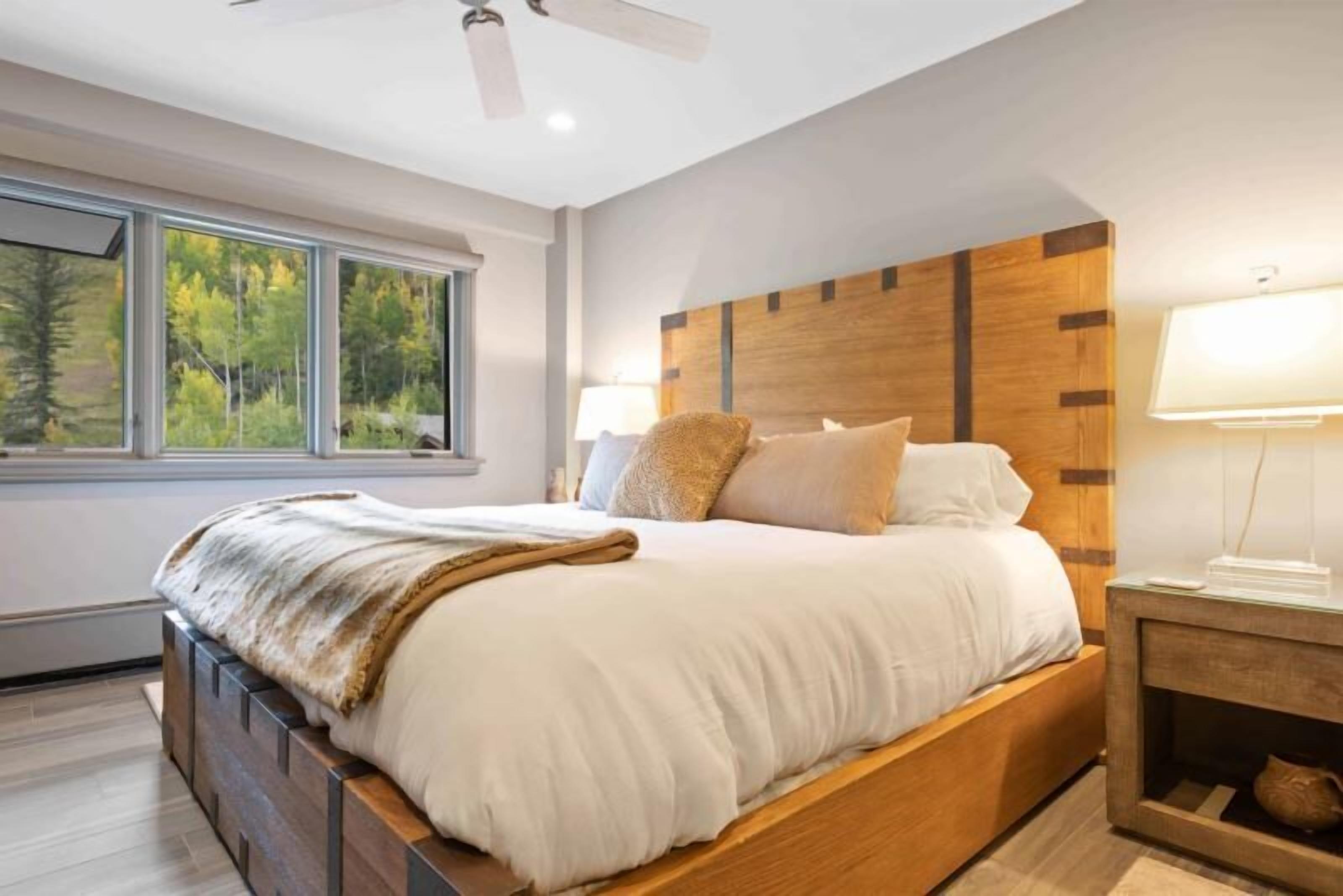 A modern bedroom with a large bed, wooden headboard, and a view of trees through the window.