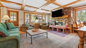 A spacious living room with a high ceiling, wooden beams, a stone fireplace, comfortable seating, and large windows.
