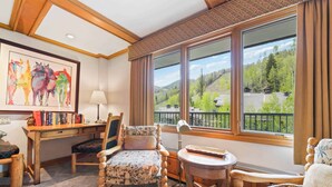 A cozy room with a wooden desk, two upholstered chairs, a small table, and a large window offering a scenic view of trees and mountains.