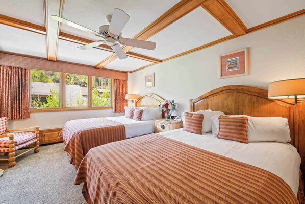 Habitación de hotel con dos camas, un ventilador de techo, una ventana con cortinas, un cabecero de madera y una mesita de noche con lámpara.