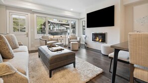 A modern living room with a fireplace, a large TV, and a view of a residential building. - Lodge Tower 278 1BR at the base of Vail Mountain (Vail)