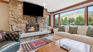 Ein Wohnzimmer mit Kamin aus Stein, einem Flachbildfernseher, einer Couch und Blick auf eine Berglandschaft.