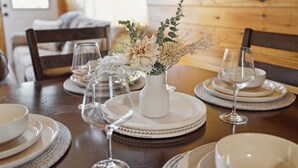 Una mesa de comedor dispuesta con platos, tazones y copas de vino, junto a un jarrón con flores secas.