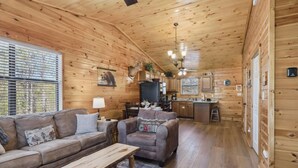Interior de una cabaña con detalles de madera, un sofá, una silla, una mesa de comedor y una zona de cocina.