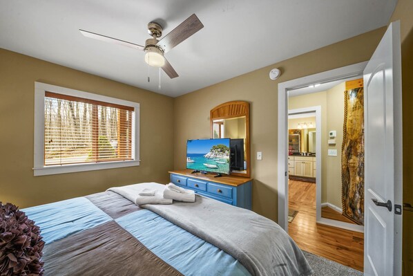 Une chambre à coucher avec un lit, un ventilateur de plafond, une télévision et une vue sur la cuisine par une porte ouverte.
