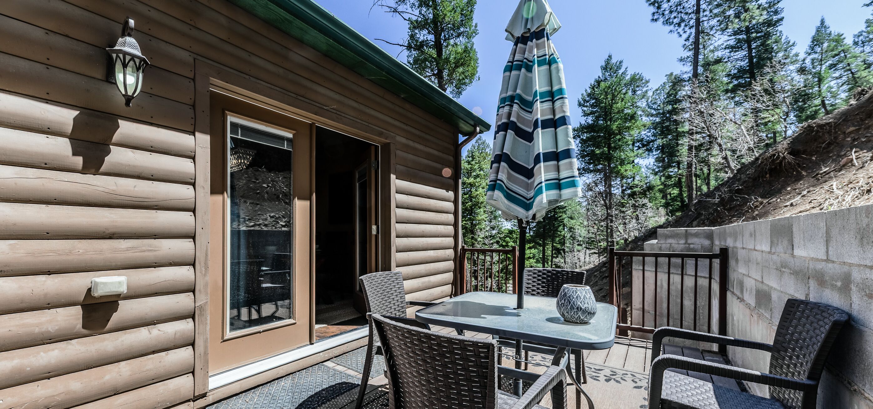 Une cabane en rondins dotée d’un patio comprenant des fauteuils en osier, une table et un parasol à rayures.
