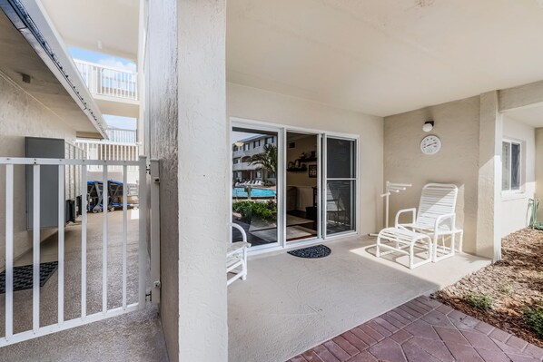 A covered outdoor area with a sliding glass door leading to a pool, white chairs, and a clock on the wall.