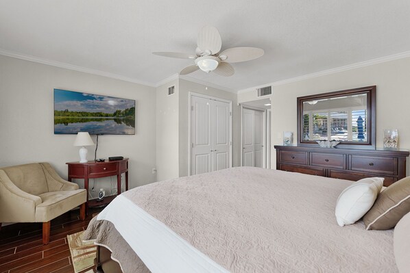 A bedroom with a bed, a chair, a desk, a dresser, a ceiling fan, and a large window.